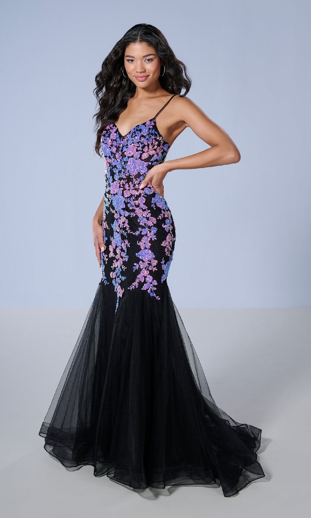 A woman wears the Tiffany 16240 long prom dress, a fitted black mermaid gown with purple and pink floral sequin appliqué, thin straps, and a tulle skirt, standing against a light gray background.